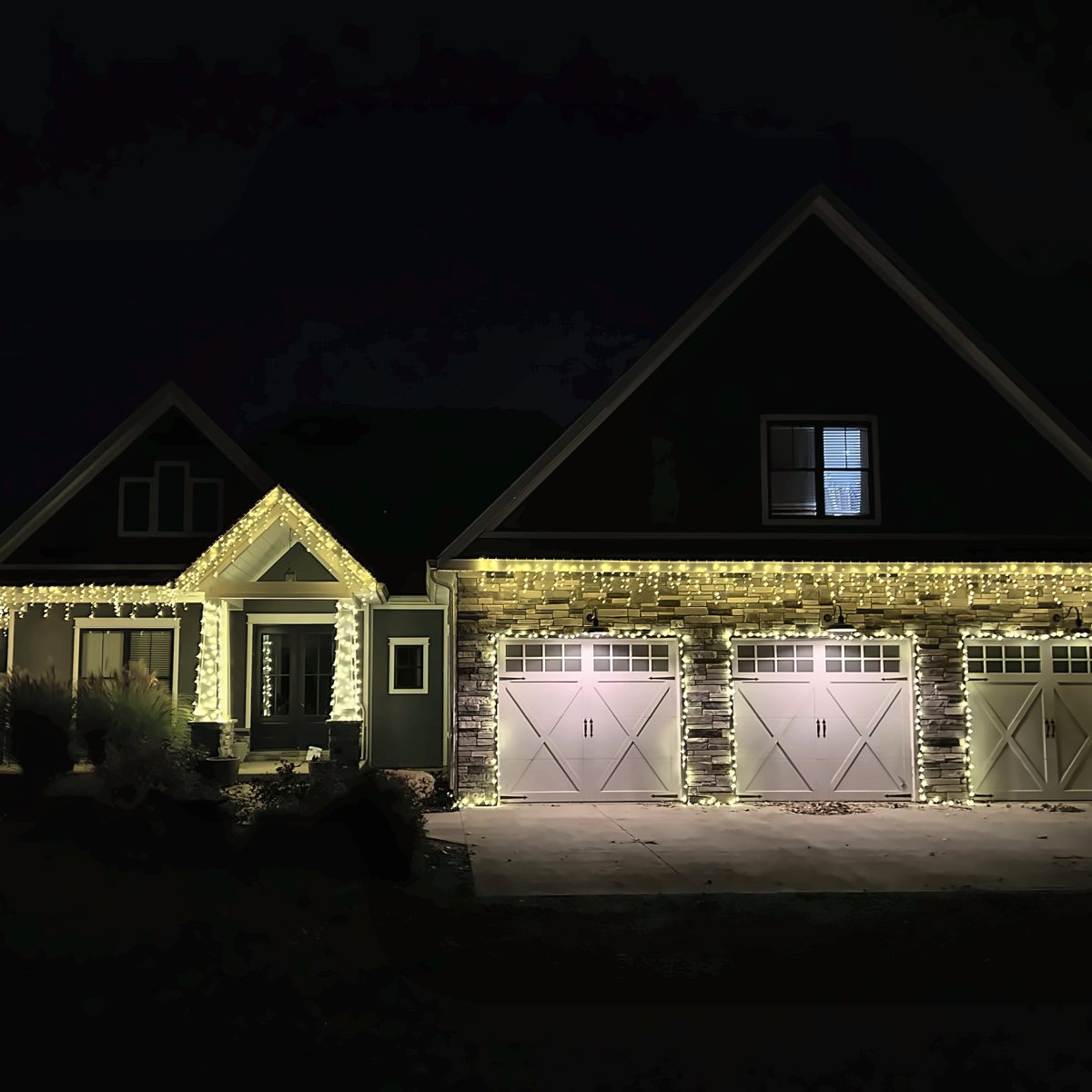 Craftsman home with warm white lights along the roofline, porch columns, and three outlined garage doors