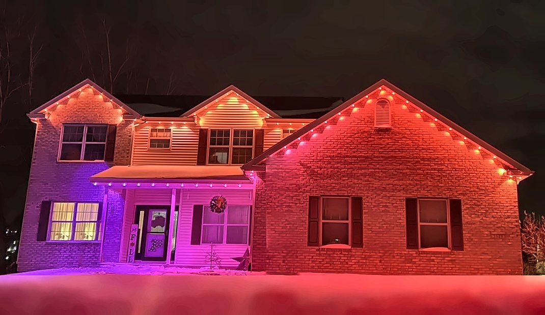 Two-story brick home with permanent lights glowing red, pink, and purple under the eaves on a snowy night